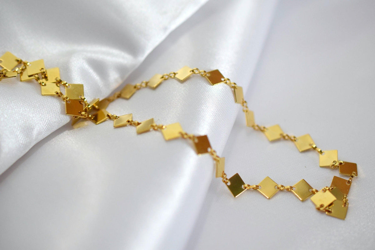 A gold necklace with diamond-shaped pendants is displayed on a white fabric surface.