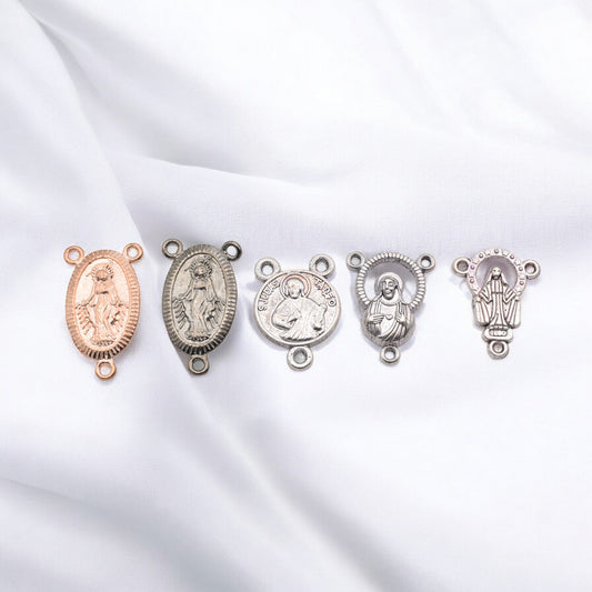 Five religious medal connectors in gold and silver tones on white fabric.