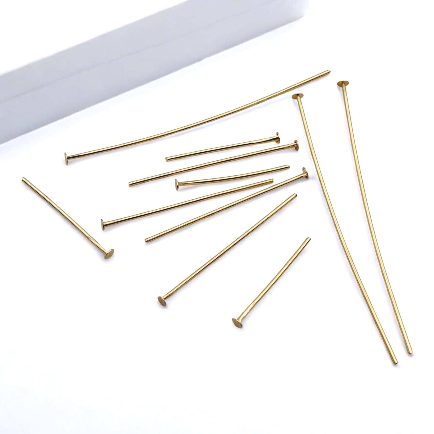 A macro photograph focusing on the base of several gold head pins. The shot emphasizes the flat, circular "nail head" design that prevents beads from sliding off the bottom of the wire.