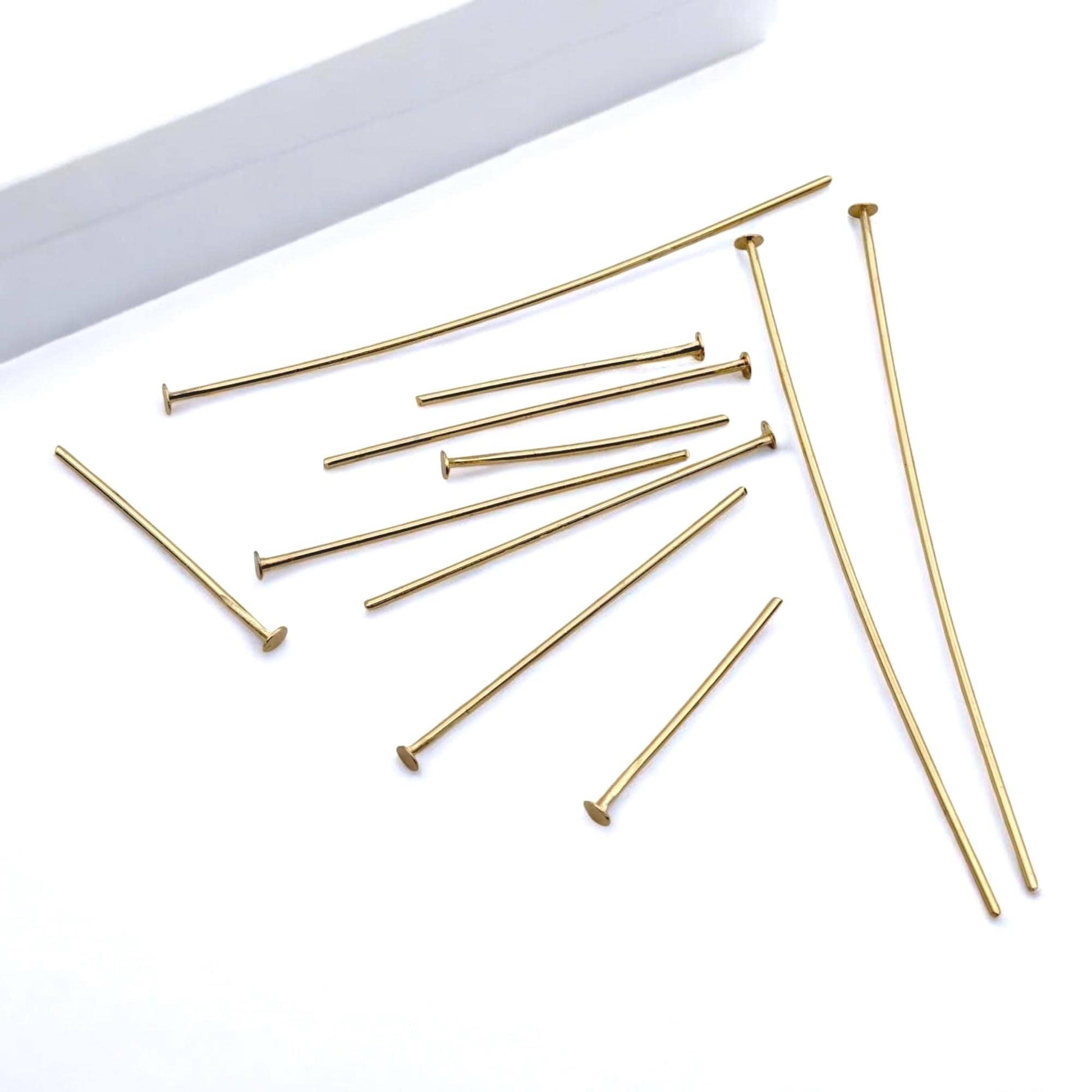 A macro photograph focusing on the base of several gold head pins. The shot emphasizes the flat, circular "nail head" design that prevents beads from sliding off the bottom of the wire.