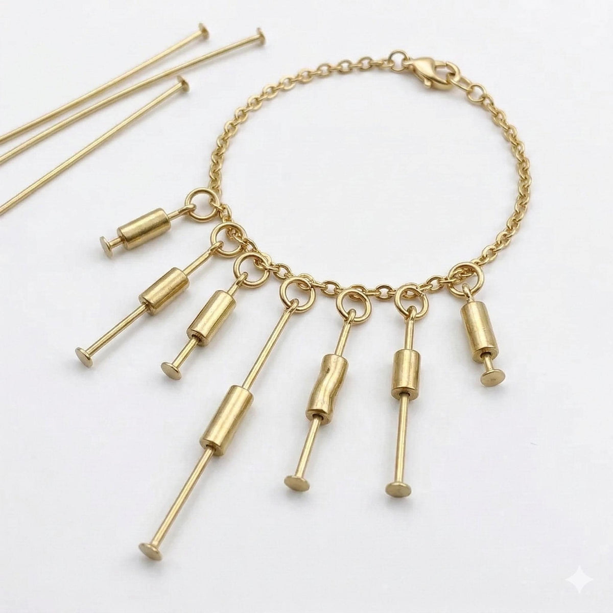 A close-up of a gold chain necklace or bracelet featuring several small gold charms attached using flat head pins. Three loose gold head pins are positioned next to the jewelry, demonstrating how the pins are used to secure beads and dangles to a link chain.