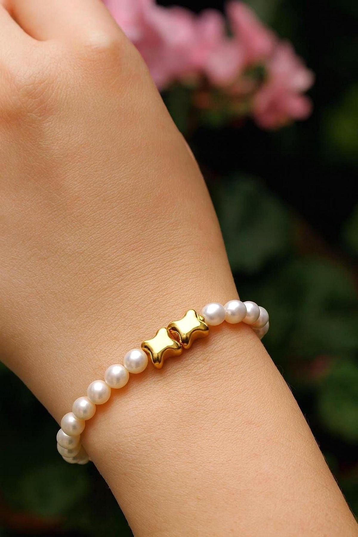 A gold-colored bracelet with white pearls is worn on a person's wrist, set against a blurred background of pink flowers.