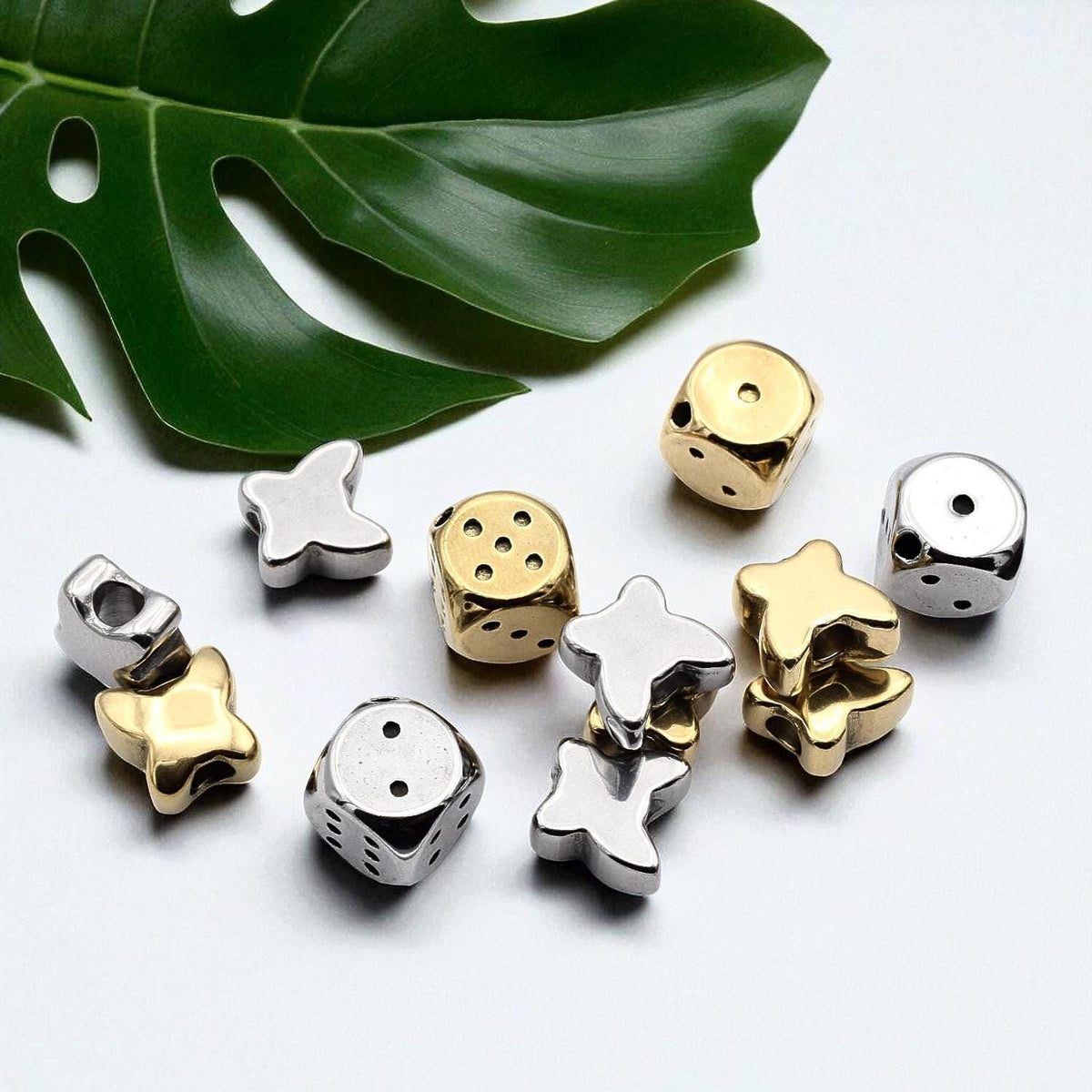 a collection of various metal beads, including dice and star-shaped beads, arranged on a white surface alongside a large green leaf.