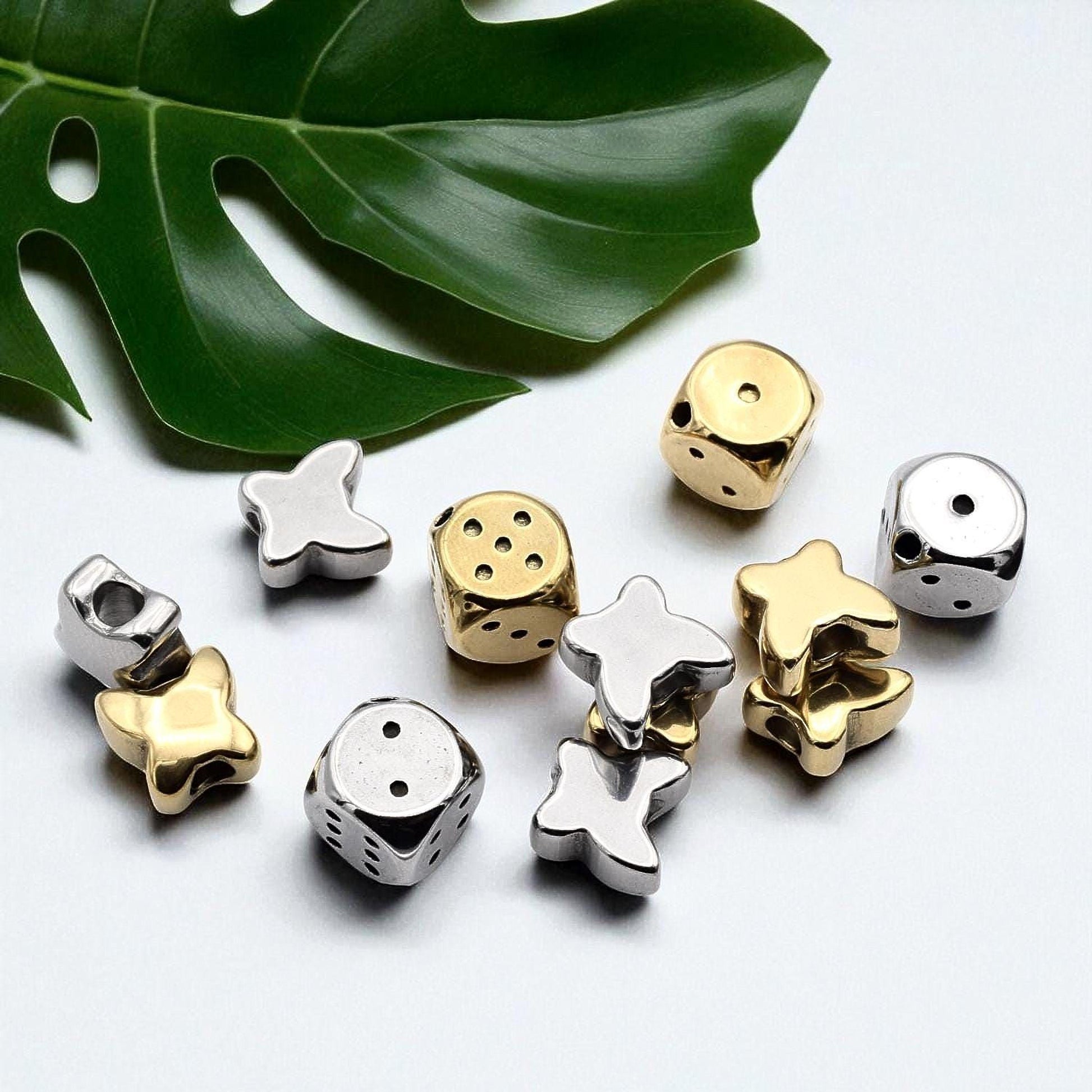 a collection of various metal beads, including dice and star-shaped beads, arranged on a white surface alongside a large green leaf.