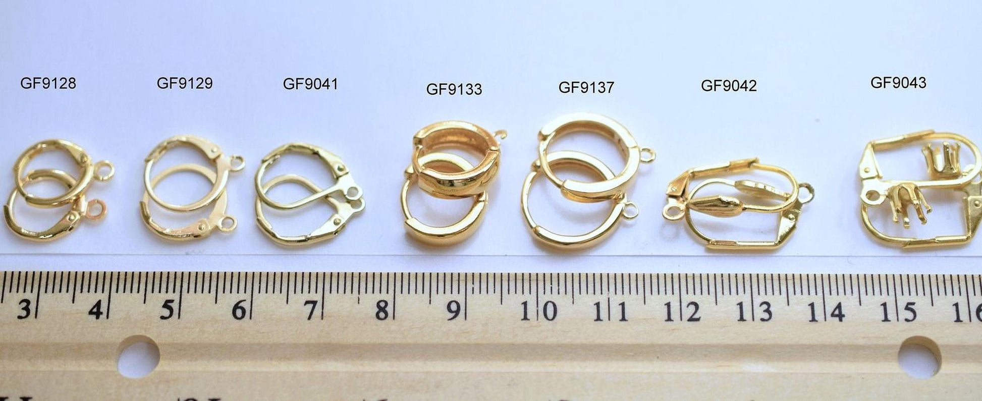 a collection of gold-colored metal pieces, including earrings and clasps, arranged on a ruler for size comparison.