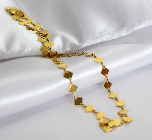 A gold necklace with diamond-shaped pendants is displayed on a white fabric surface.